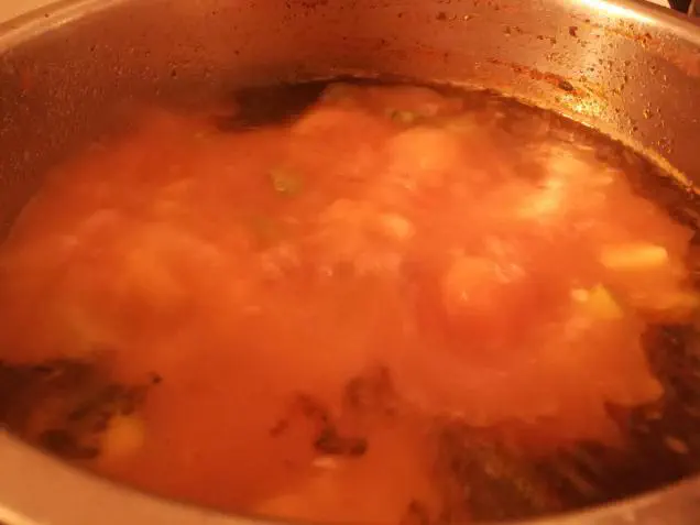 A blurry boiling pot of red soup with some chunks of zucchinis and a layer of darker red seasonings floating on top of the liquid
